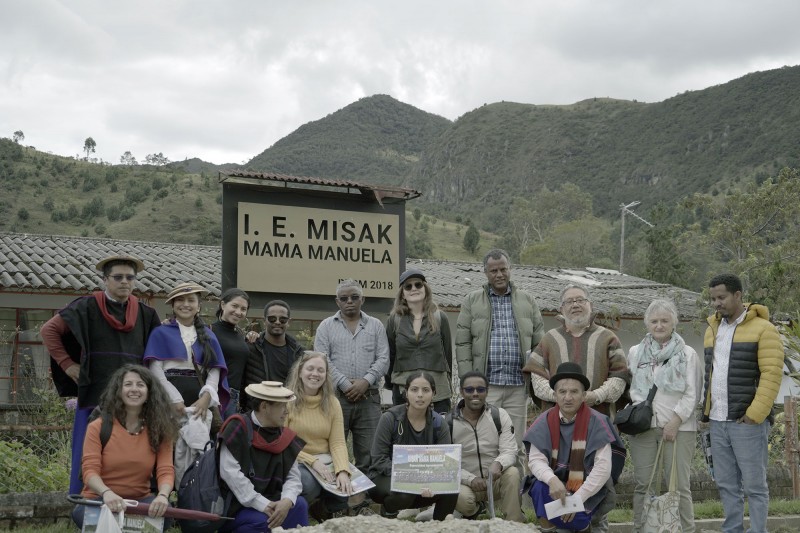 Smiling Hub researchers and members of the Misak community wearing traditional dress sit and stand together in front of a sign that reads 'I.E. Misak Mama Manuela'